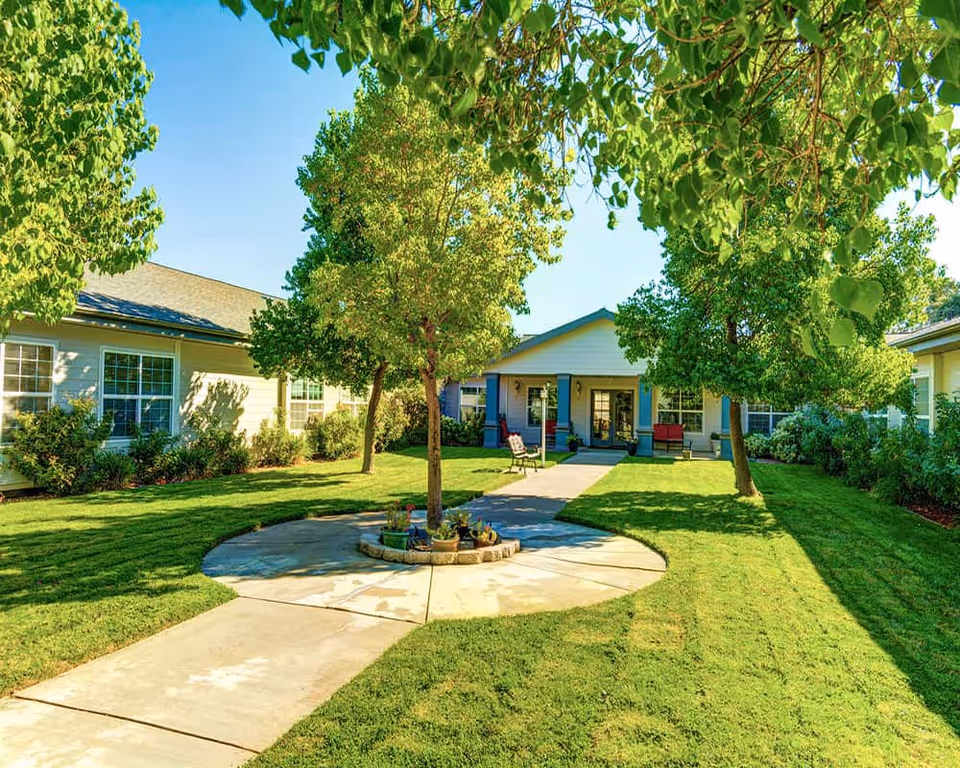 Sunlit courtyard with a paved walkway and trees leading to the front entrance of a single-story assisted living building.
