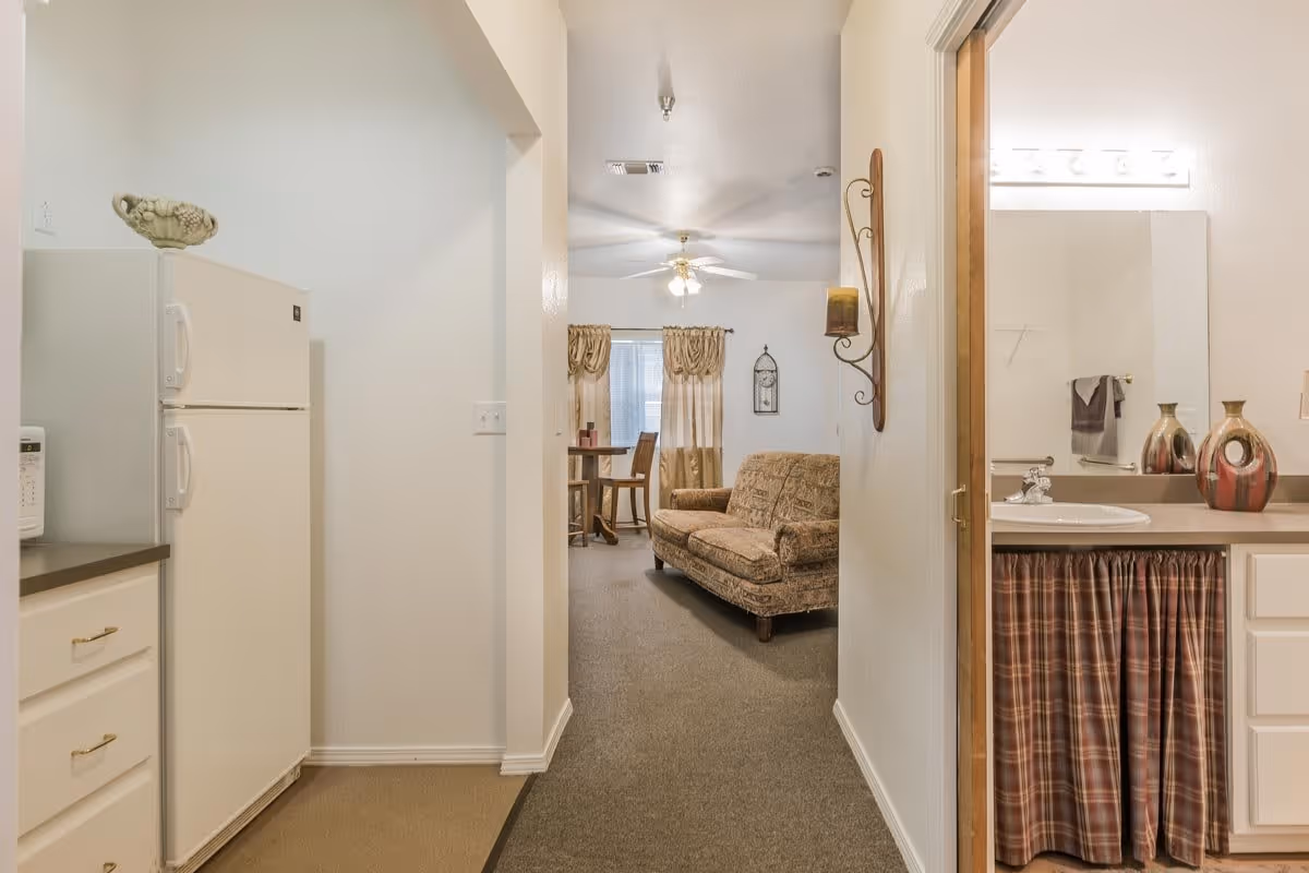 View down a hallway in a senior living facility showing a small kitchen area with a white refrigerator and microwave on the left, a living room with a patterned sofa and a small dining table with chairs in the background, and a bathroom vanity with a sink, mirror, and decorative vases on the right.