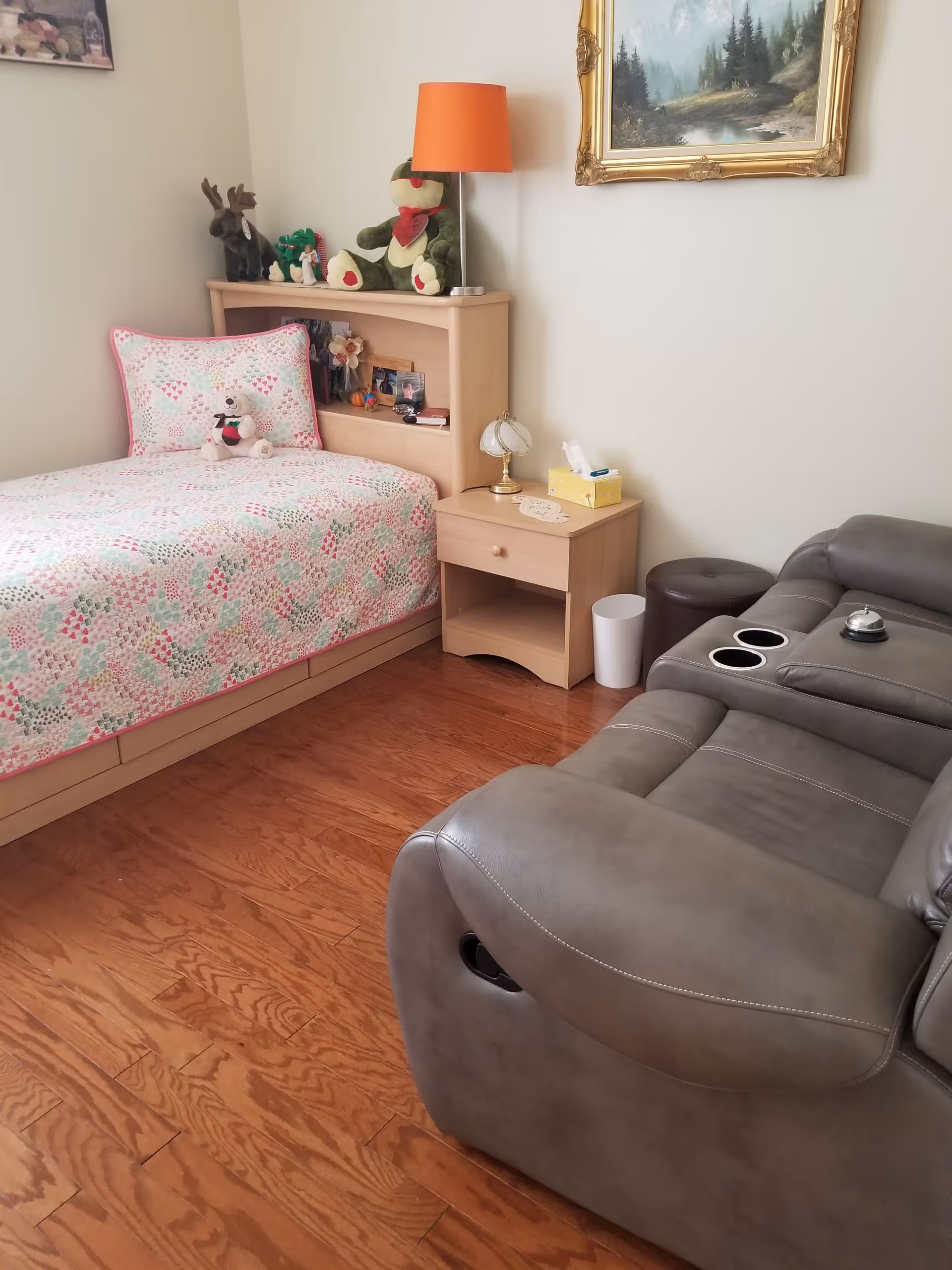 A cozy bedroom with a single bed covered in a pastel-colored quilt and a teddy bear on the pillow. Next to the bed is a wooden nightstand with a small lamp and a tissue box. On the headboard shelf are several stuffed animals and small decorative items. A gray recliner chair with cup holders and a service bell is positioned near the nightstand. The room has wooden flooring and a framed landscape painting on the wall.