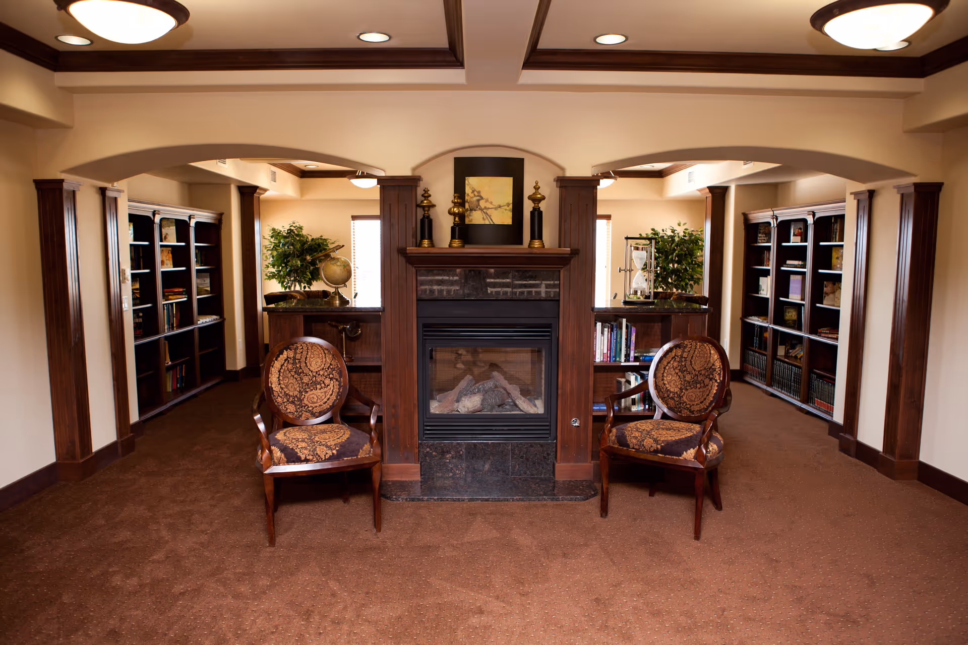A cozy interior common area featuring a central fireplace with decorative items on the mantel. Two ornate wooden chairs with patterned upholstery are placed on either side of the fireplace. The room has warm lighting, carpeted floors, and built-in bookshelves filled with books and decorative objects along the walls. There are also potted plants adding greenery to the space.