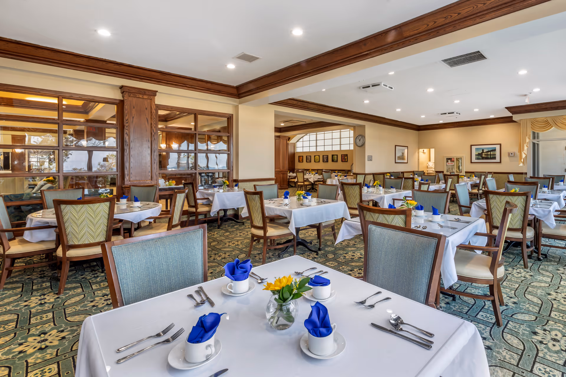 A spacious dining room with multiple tables covered in white tablecloths, each set with cups, silverware, and blue folded napkins. The room features wooden trim, patterned carpet, and large windows with a view of an adjacent room. Yellow flowers in small vases decorate each table.