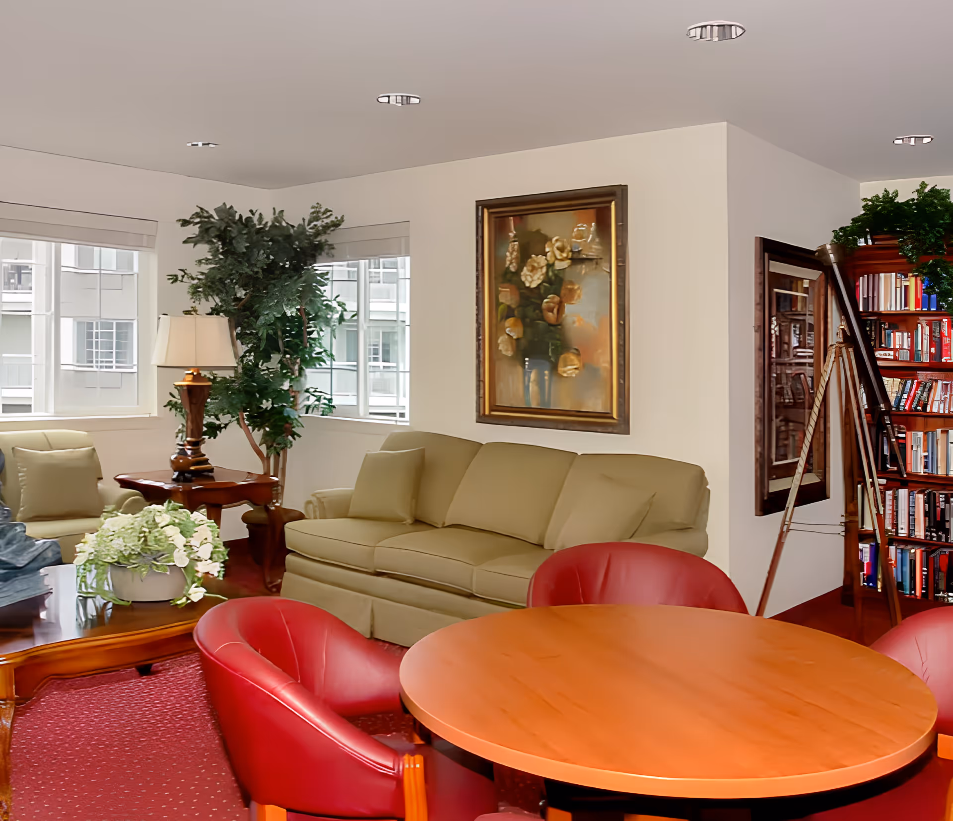 Bright common living room with a sofa, round wooden table surrounded by red chairs, coffee table, potted plants, a bookshelf and a framed painting.