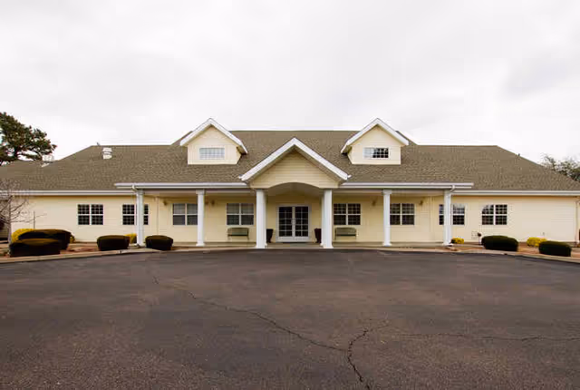 Single-story cream-colored building with a central covered entrance and columns facing a paved parking area under an overcast sky.