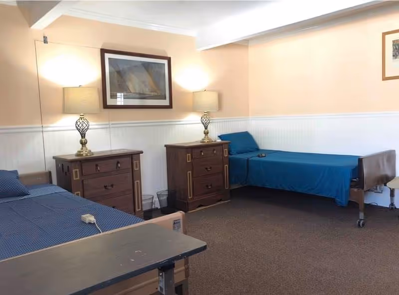 Two single beds with blue bedding, wooden nightstands and lamps in an assisted living bedroom.