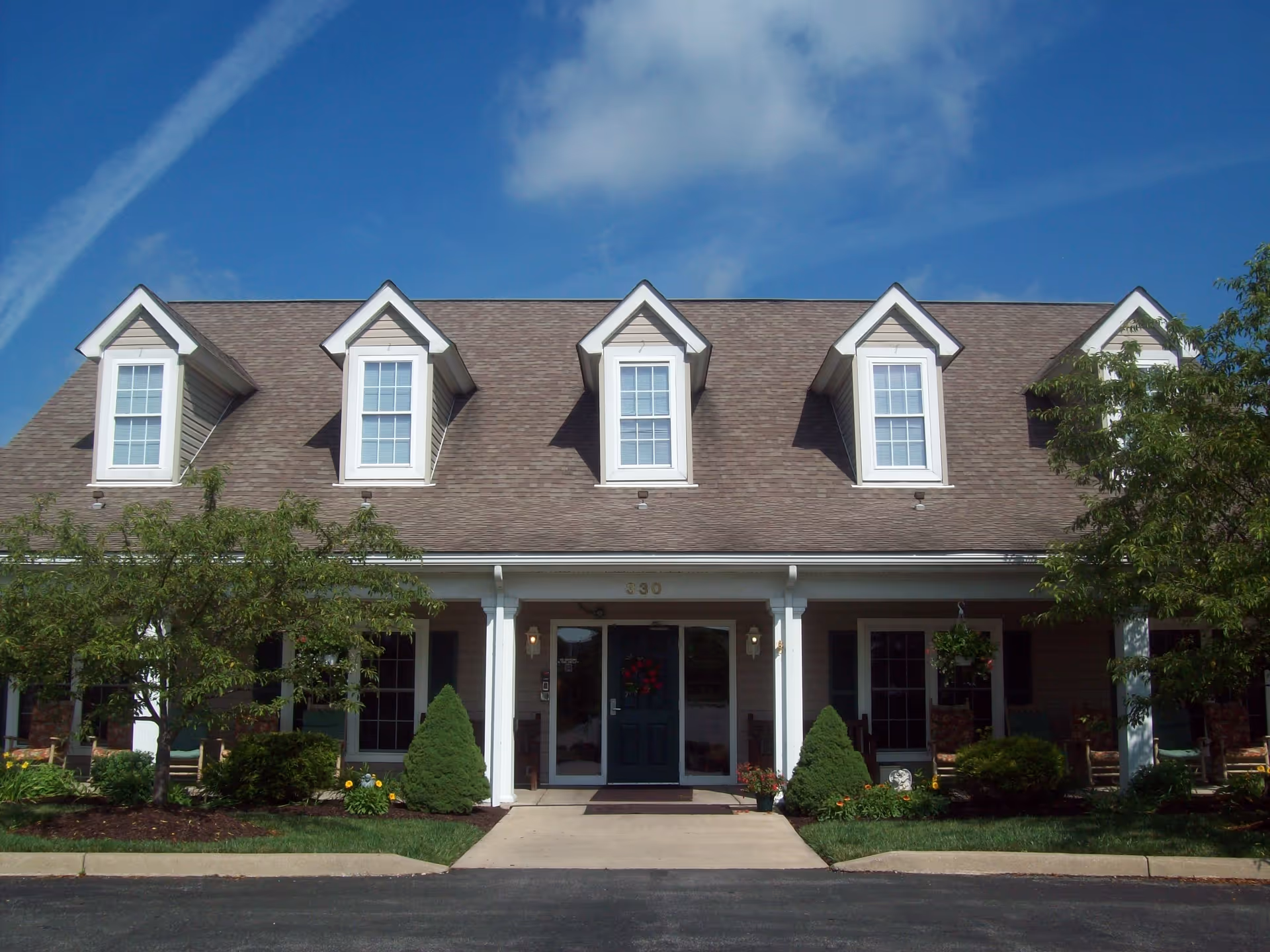 Front exterior view of a two-story building with a brown shingled roof and four dormer windows. The entrance has double glass doors with a wreath, flanked by white columns and outdoor lights. There are shrubs, small trees, and a well-maintained lawn in front of the building under a blue sky with some clouds.
