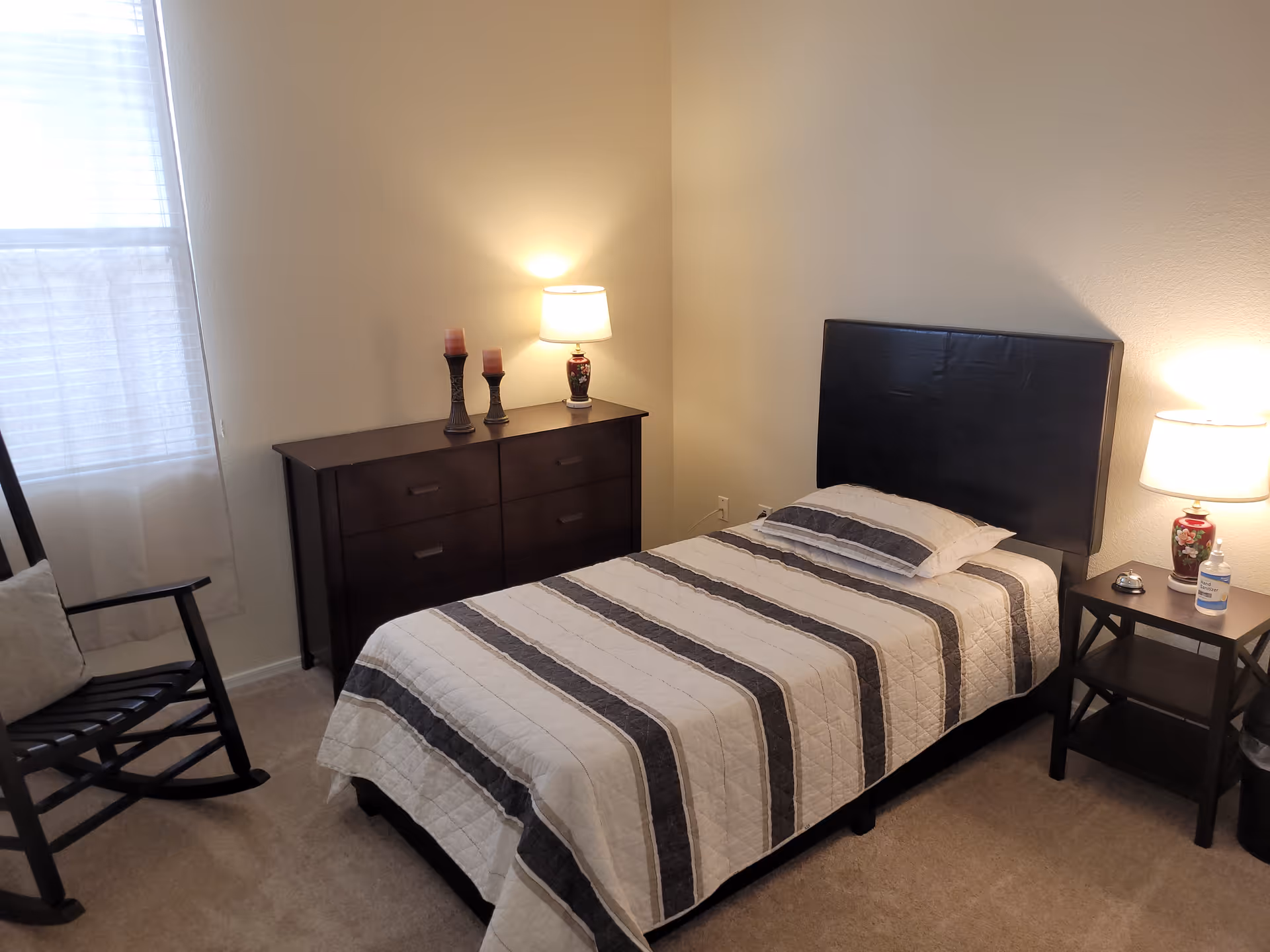 A small bedroom with a single bed covered in a striped quilt. There is a black headboard, a nightstand with a lamp, hand sanitizer, and a bell. A dresser with two candles and a lamp is against the wall. A rocking chair with a cushion is near a window with sheer white curtains.