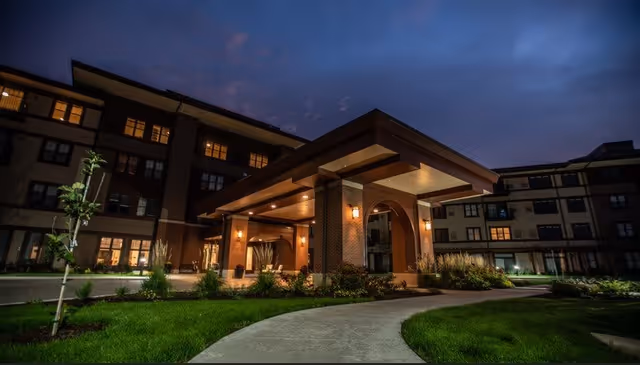 Exterior view of a senior living facility at dusk with a well-lit entrance canopy, surrounded by landscaped greenery and a curved walkway leading to the entrance.