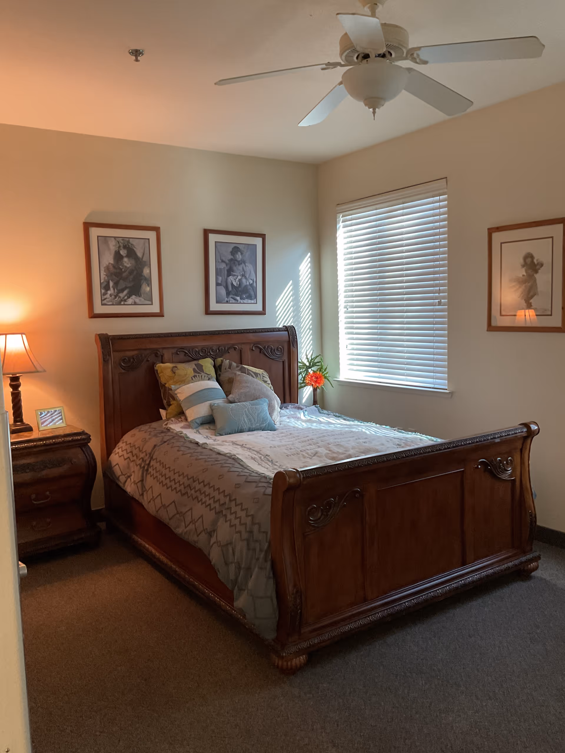 A cozy bedroom with a wooden bed frame and headboard, neatly made bed with multiple pillows, a nightstand with a lamp and a small picture frame, three framed pictures on the walls, a window with closed blinds, and a ceiling fan above.
