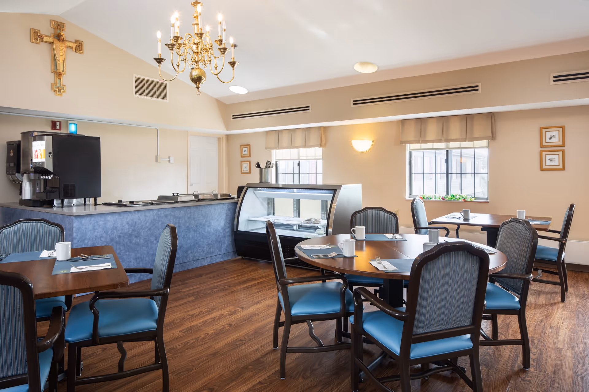 A dining room in Vincentian Home with wooden floors, round and square tables set with placemats, utensils, and white mugs. The room features blue cushioned chairs, a chandelier hanging from the ceiling, a beverage dispenser, and a display case. There are windows with beige valances and framed pictures on the walls.