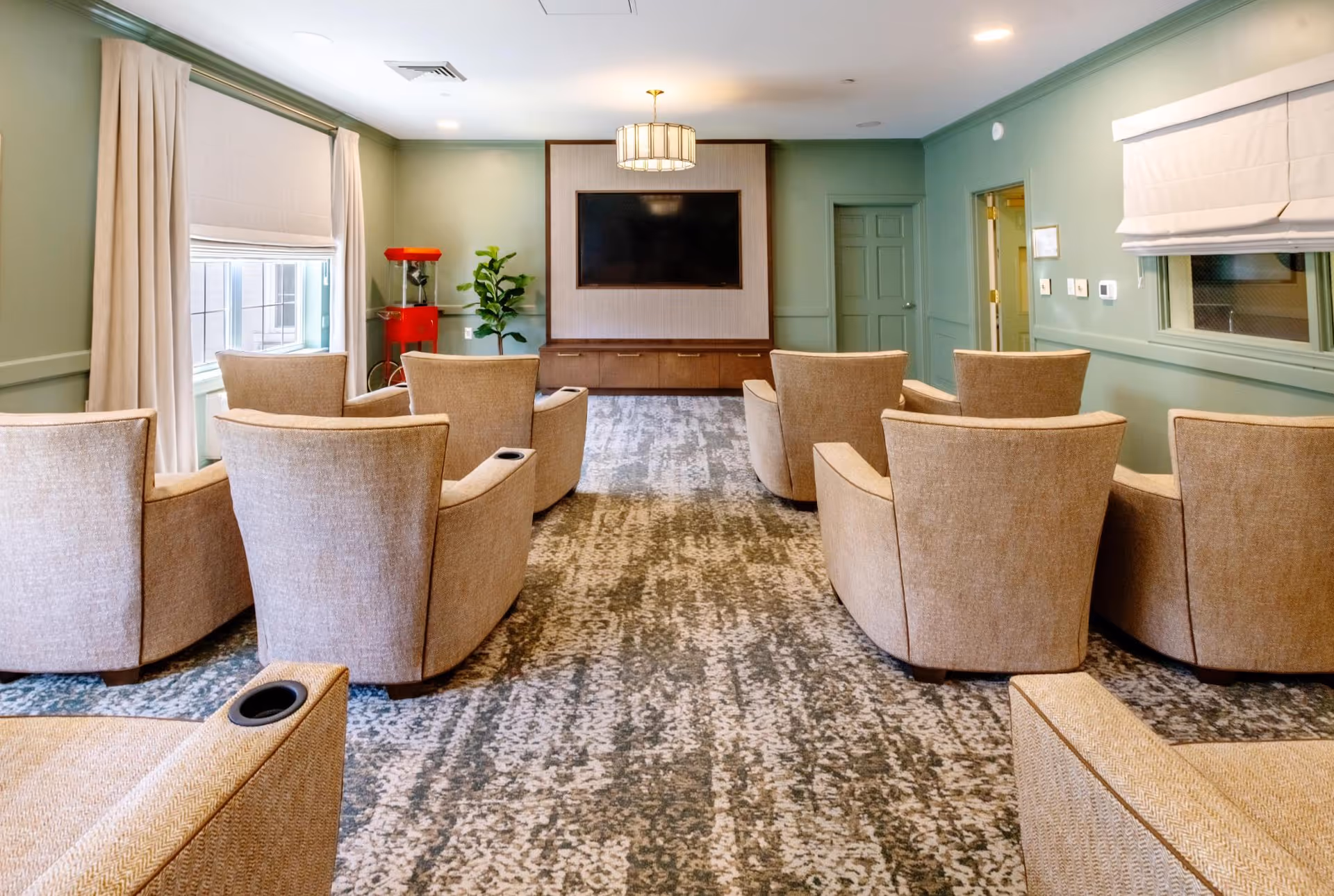 A cozy media room with eight beige armchairs arranged in two rows facing a large flat-screen TV mounted on a wooden panel wall. The room has light green walls, a patterned carpet, a red popcorn machine in the corner, a potted plant, and windows with white roman shades and beige curtains.