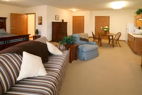 A spacious senior living apartment interior featuring a striped sofa with white pillows, a blue armchair, a wooden side table with a plant, a wooden dining table with four chairs, and a kitchenette with wooden cabinets and appliances. The room has beige carpeting and multiple wooden doors leading to other rooms.