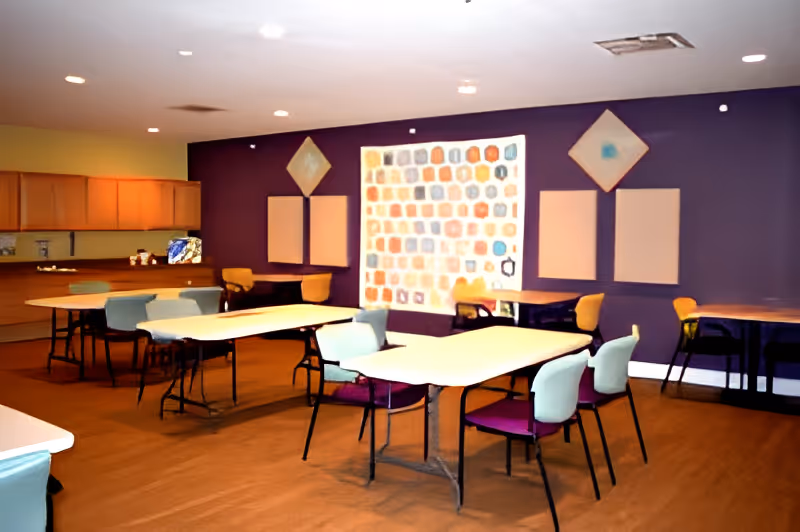 A room with several tables and chairs arranged for group seating, featuring a purple accent wall with abstract artwork and a kitchen area with wooden cabinets in the background.
