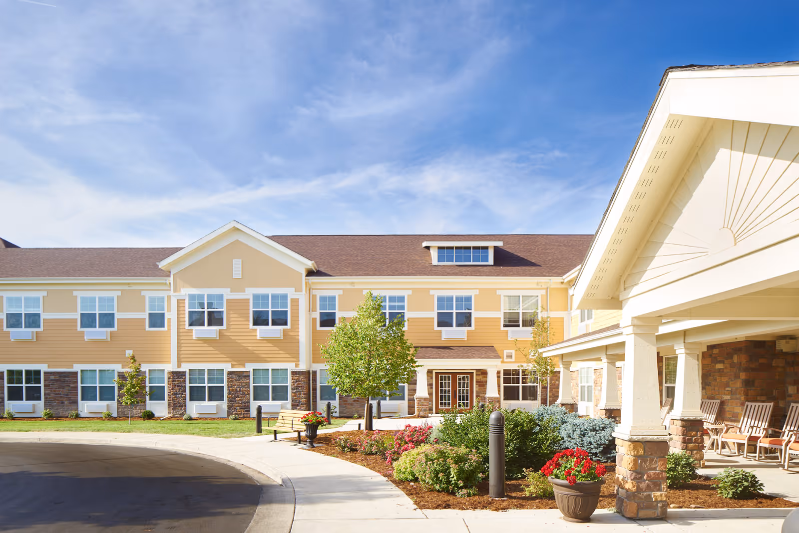 Exterior view of a senior care community building with beige and yellow siding, multiple windows, a covered entrance with columns, landscaped garden with flowers and shrubs, a bench, and a clear blue sky.