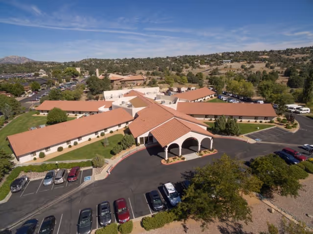 Aerial view of Granite Creek Health and Rehabilitation facility showing a large building with a red-tiled roof and multiple wings extending outward. The building is surrounded by parking lots with several parked cars, landscaped greenery, and trees. The background features rolling hills and a partly cloudy blue sky.