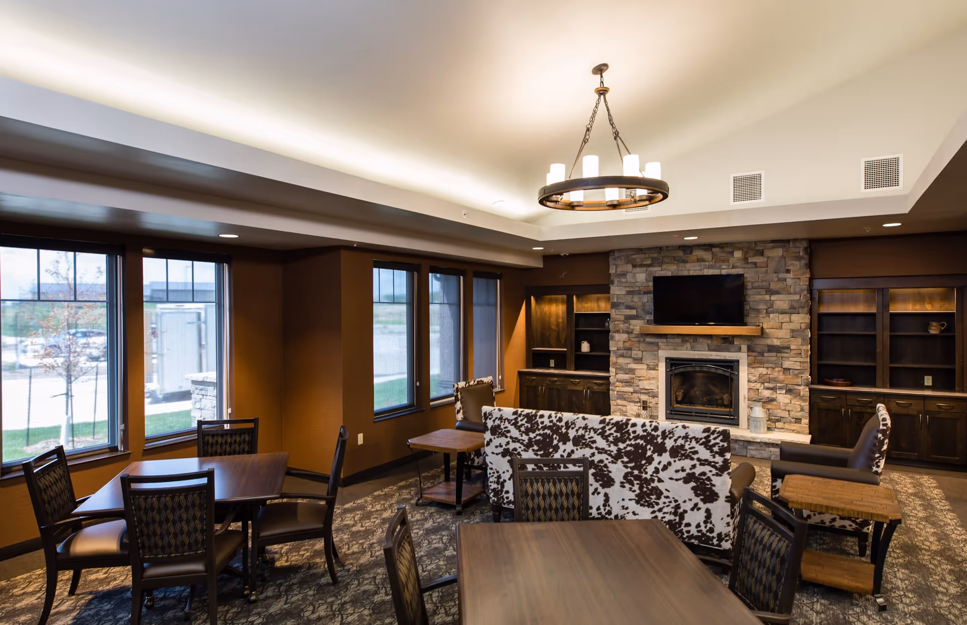 A cozy living room area in an assisted living facility featuring a stone fireplace with a mounted TV above it, built-in wooden shelves on either side, multiple tables and chairs, and a sofa with a black and white patterned upholstery. Large windows allow natural light to enter the room.