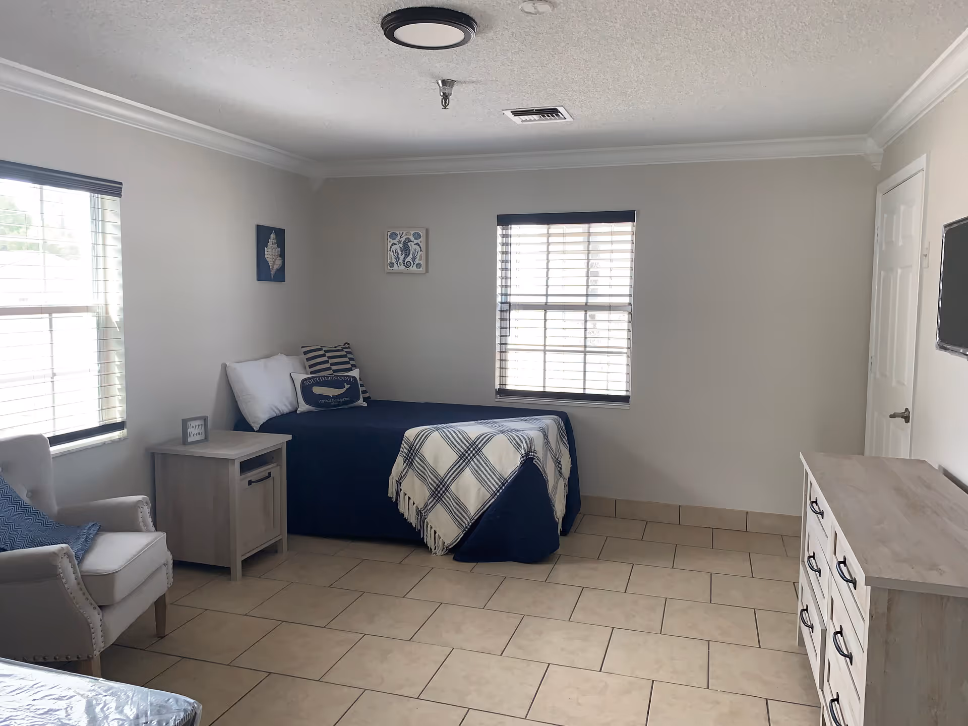 Simple single-occupancy bedroom with a single bed, nightstand, armchair, dresser, and two windows.