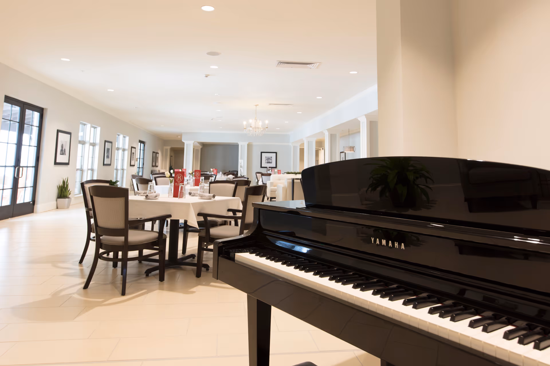 A bright dining room with set tables and chairs and a black Yamaha piano in the foreground.