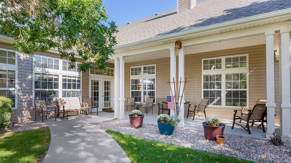 Covered outdoor patio with chairs, tables, and potted plants along the front of a senior living building.