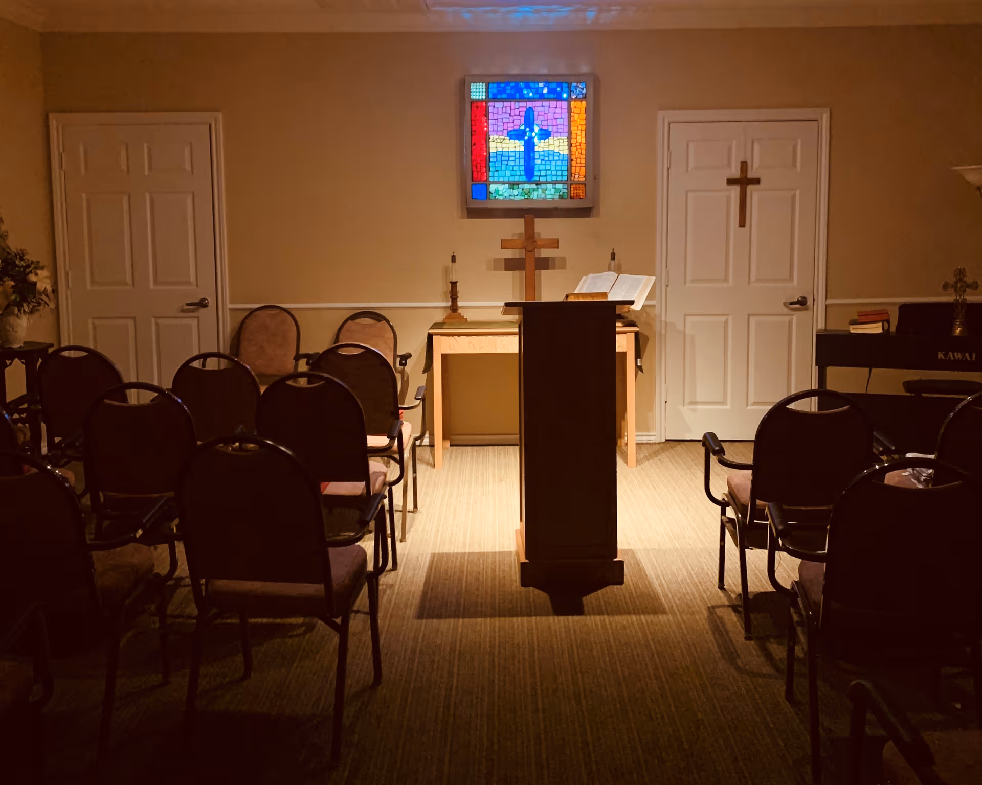 Small chapel interior with rows of chairs, a central lectern, an altar, and a stained-glass cross on the wall.