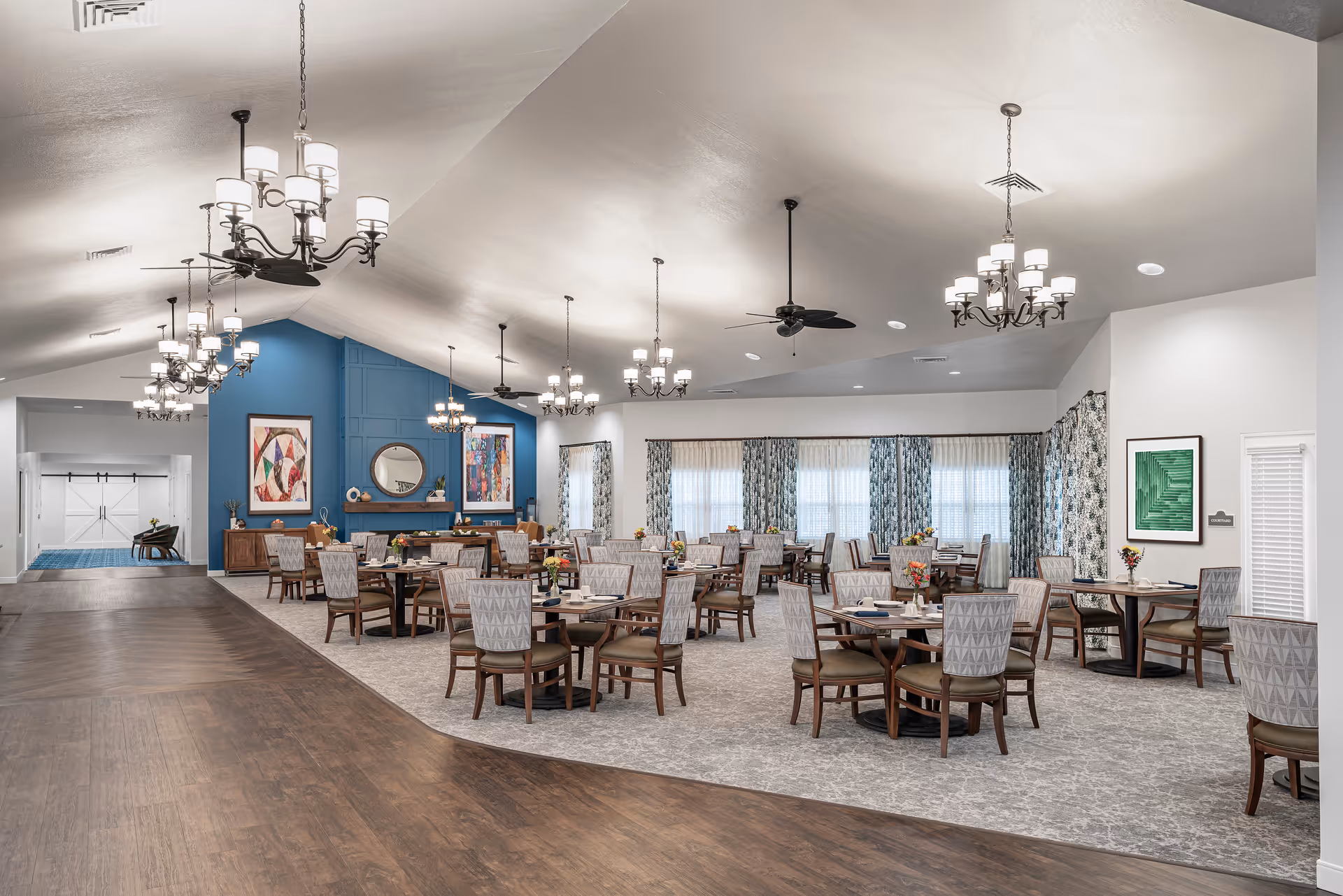 A spacious dining room with multiple round tables and chairs arranged neatly. The room features a high vaulted ceiling with several chandeliers and ceiling fans. The walls are decorated with framed artwork and a large blue accent wall with a round mirror. Large windows with patterned curtains allow natural light to fill the space. The floor transitions from wood to carpet in the dining area.
