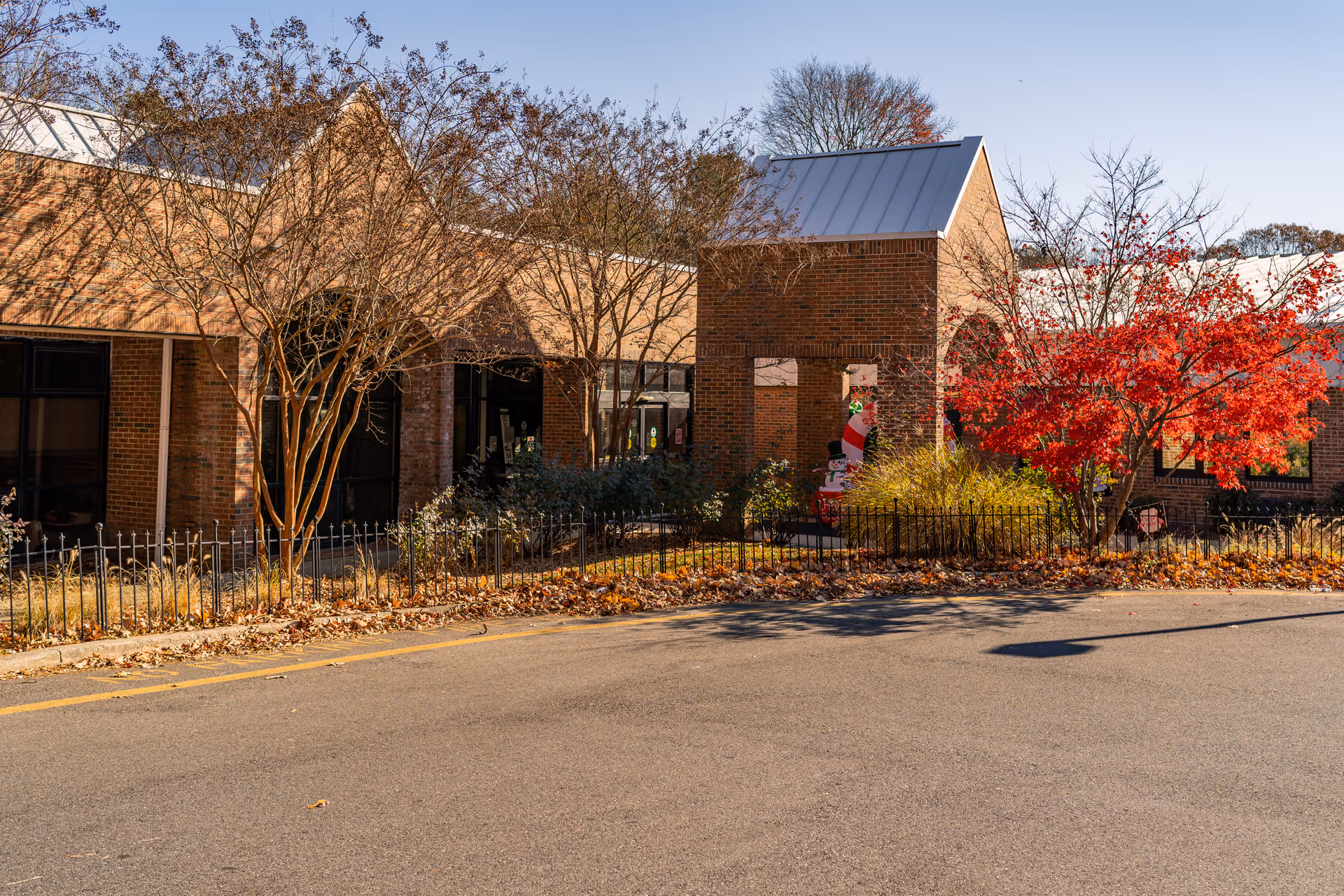 Exterior view of a brick building with a metal roof, surrounded by trees with autumn foliage including a vibrant red tree. There is a small black metal fence along the front and a paved driveway or road in the foreground. The building has large windows and an entrance area with a brick archway.