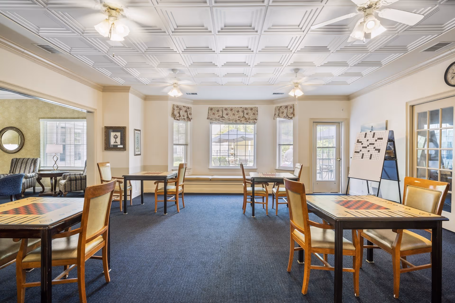A bright and spacious common room with several tables and chairs arranged for games or social activities. The tables have game boards printed on them. The room has a decorative ceiling with multiple ceiling fans and large windows with patterned valances letting in natural light. There is a large crossword puzzle board on an easel near a door leading outside, and a seating area with armchairs is visible through an open doorway.