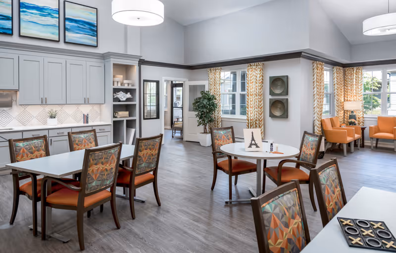 Bright communal dining and activity room with tables, patterned chairs, a kitchenette, and a seating area near windows.