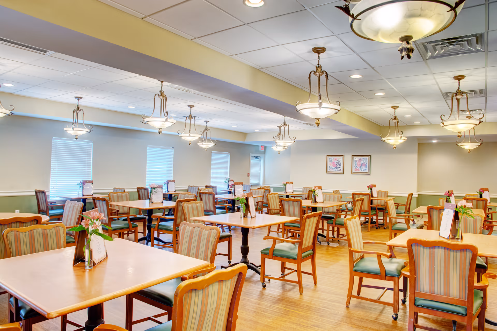 Bright dining room with multiple wooden tables and striped upholstered chairs, chandeliers overhead, and small flower centerpieces.
