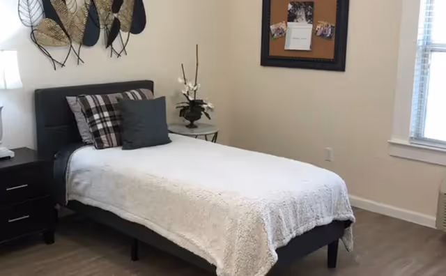 A neatly made single bed with a white blanket and two pillows, one plaid and one solid dark gray, in a bright room with light-colored walls and wood flooring. There is a black nightstand with a lamp on the left side of the bed, a small round table with a flower arrangement on the right, a decorative wall art above the bed, a bulletin board on the wall, and a window with blinds on the right side.