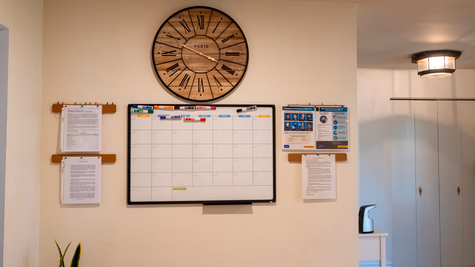Wall with a large round wooden clock showing the time as 3:40, a whiteboard calendar with colored magnets, and several pinned notices and papers. To the right, there is a small white table with a black and white automatic soap dispenser and a hallway with closed white closet doors and a ceiling light fixture.