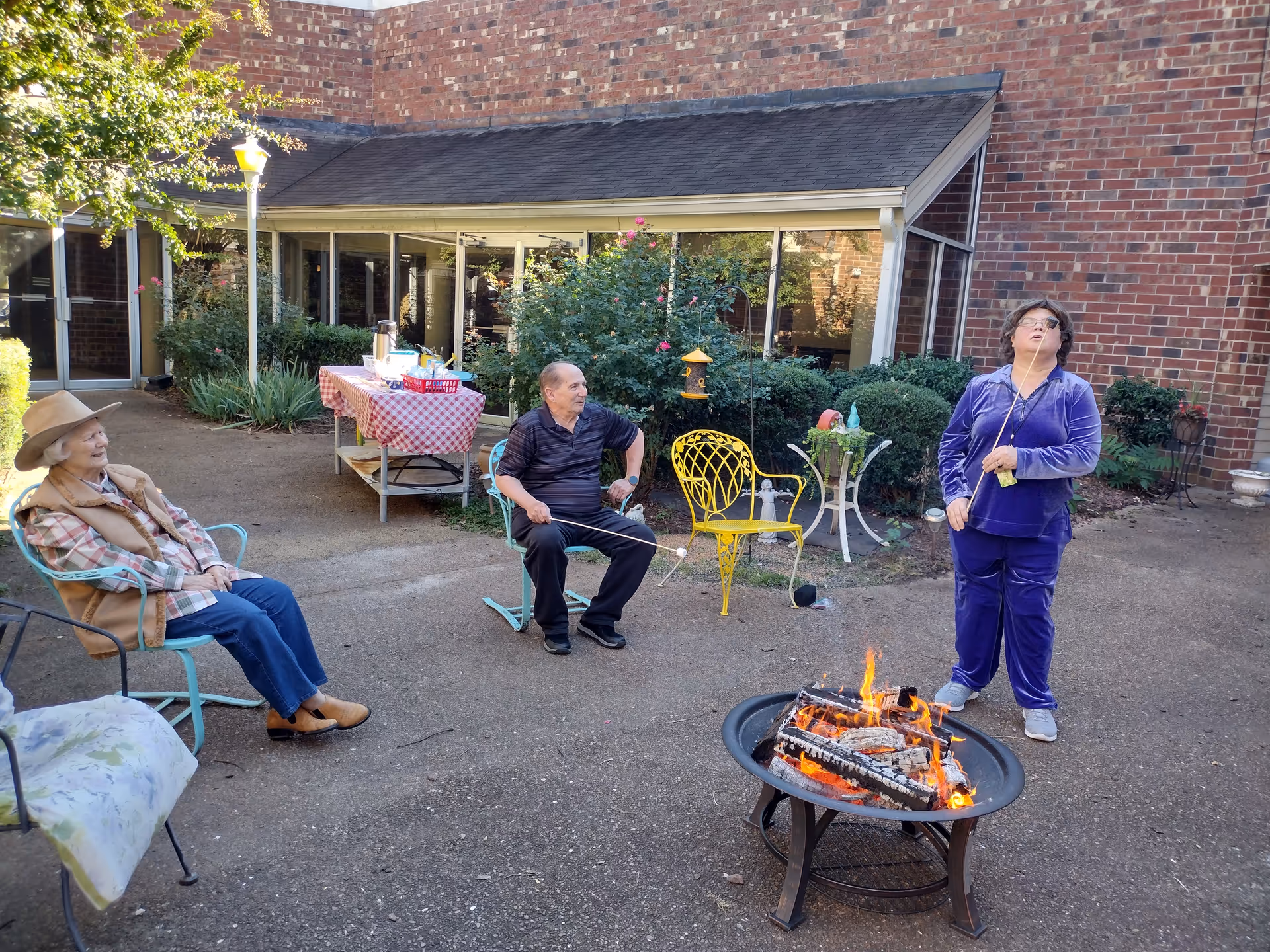 Three elderly people enjoying an outdoor fire pit in a courtyard of a senior living facility. One person is sitting on a blue metal chair wearing a hat and a vest, another person is sitting on a blue metal chair holding a stick with a marshmallow, and the third person is standing near the fire pit roasting a marshmallow. There is a table with a red and white checkered tablecloth and refreshments in the background, surrounded by bushes and brick building walls.
