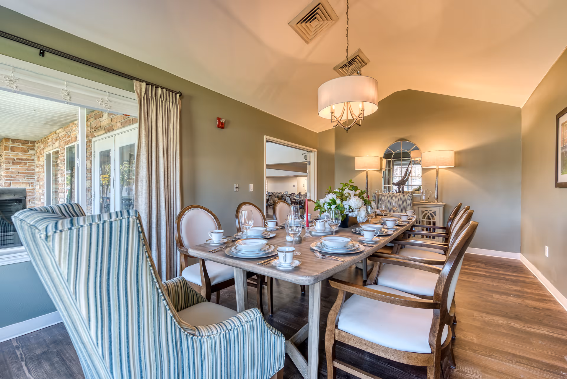 A well-lit dining room with a long wooden table set for a meal, featuring white plates, cups, and glassware. The room has a mix of upholstered chairs, including striped armchairs and wooden chairs with white cushions. There is a large window with curtains on one side, and a sideboard with two lamps and a decorative mirror on the far wall. The floor is wooden, and a modern chandelier hangs above the table.