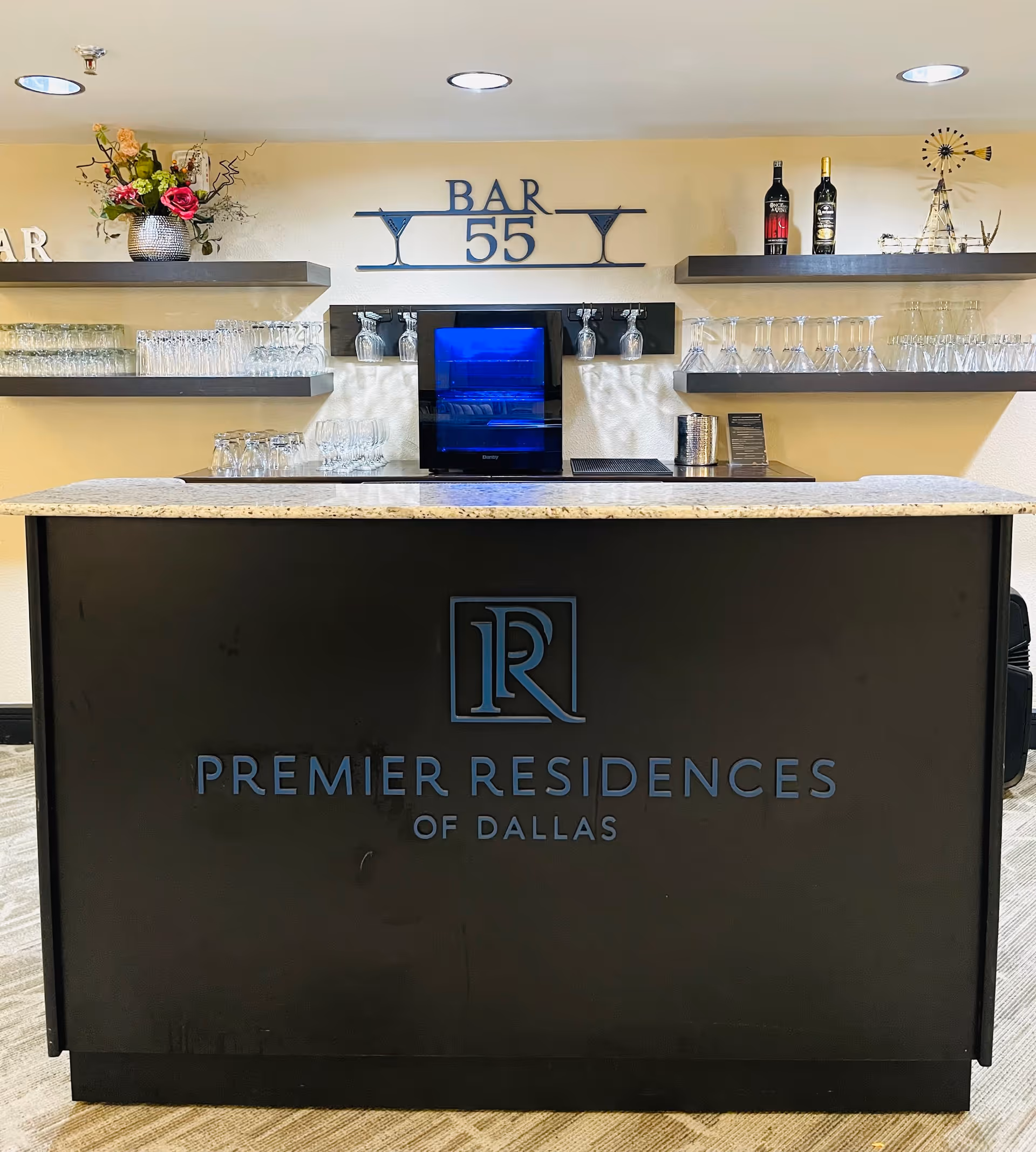A modern indoor bar area with a black counter that has the logo and text 'Premier Residences of Dallas' on the front. Behind the counter are shelves holding various glassware, two bottles of wine, a small decorative windmill, and a flower arrangement. Above the shelves is a sign that reads 'BAR 55' with martini glass icons on either side.