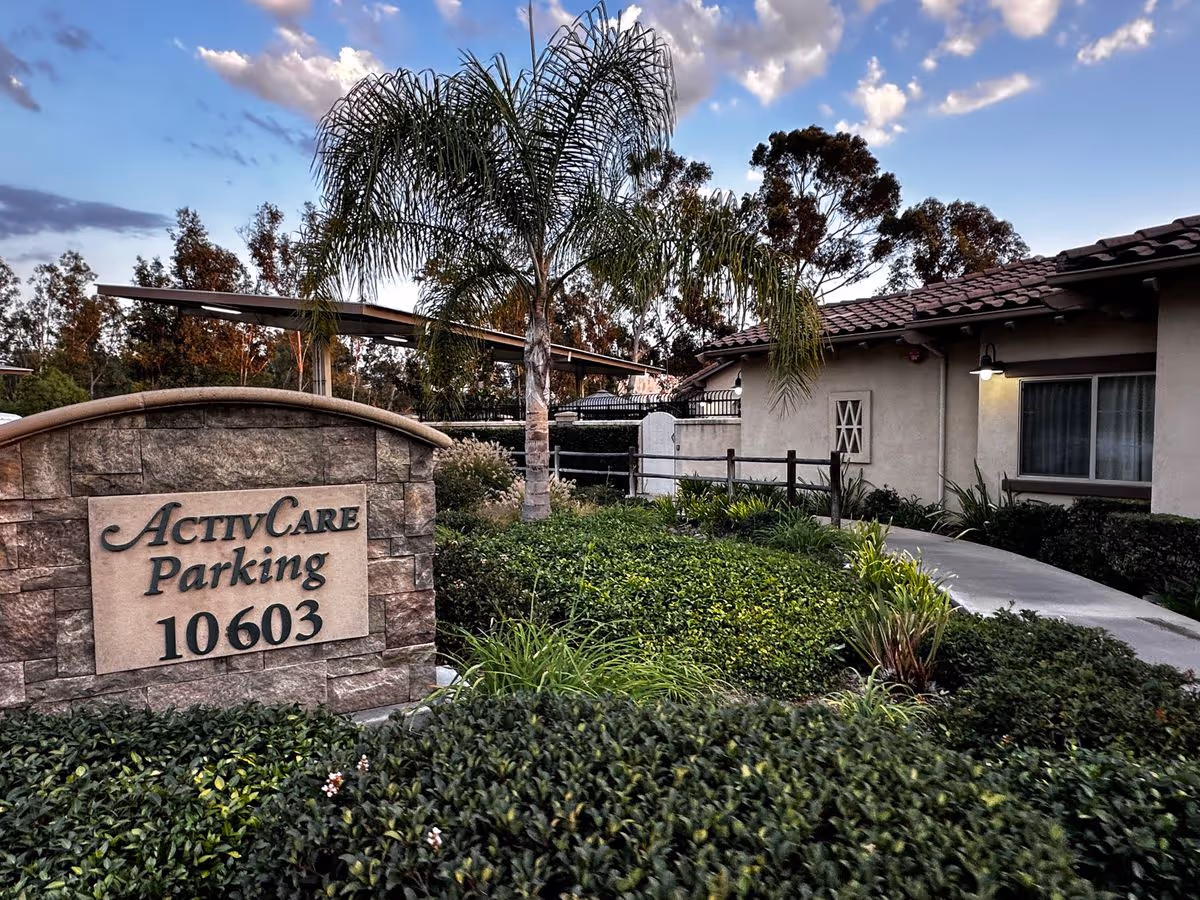 Stone sign reading "ActivCare Parking 10603" in a landscaped front yard with palm trees beside a single-story building.