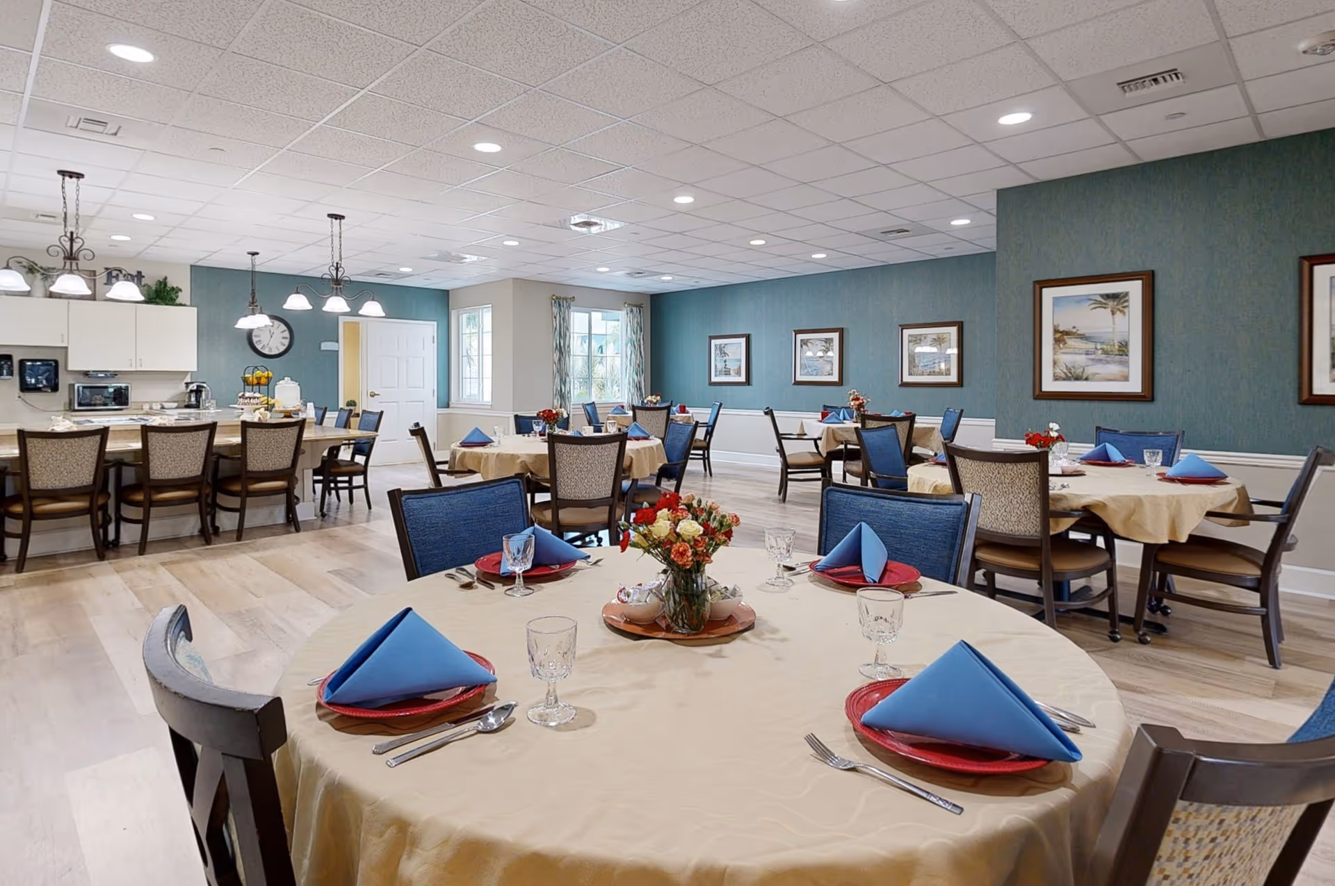 A dining room in a senior living facility with round tables covered in beige tablecloths, each set with red plates, blue folded napkins, silverware, and glasses. The room has blue walls with framed artwork, wooden chairs with blue cushions, and a kitchen area with a counter and bar stools in the background. Ceiling lights illuminate the space.