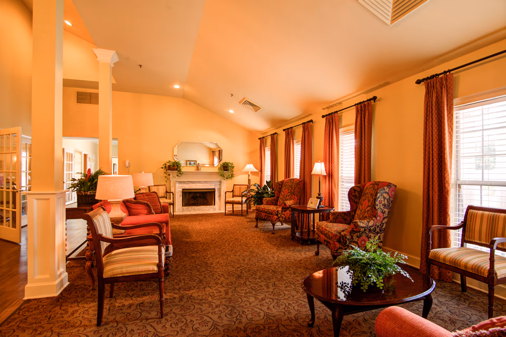 A warm and inviting living room area in a senior living facility with multiple upholstered chairs, side tables with lamps, a coffee table with a plant, large windows with red curtains, and a white fireplace with a mirror and plants on the mantel.
