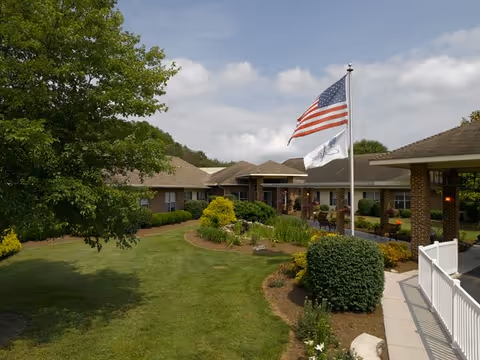 Front entrance of a single-story senior living building with landscaped lawn, flagpoles, and a walkway.