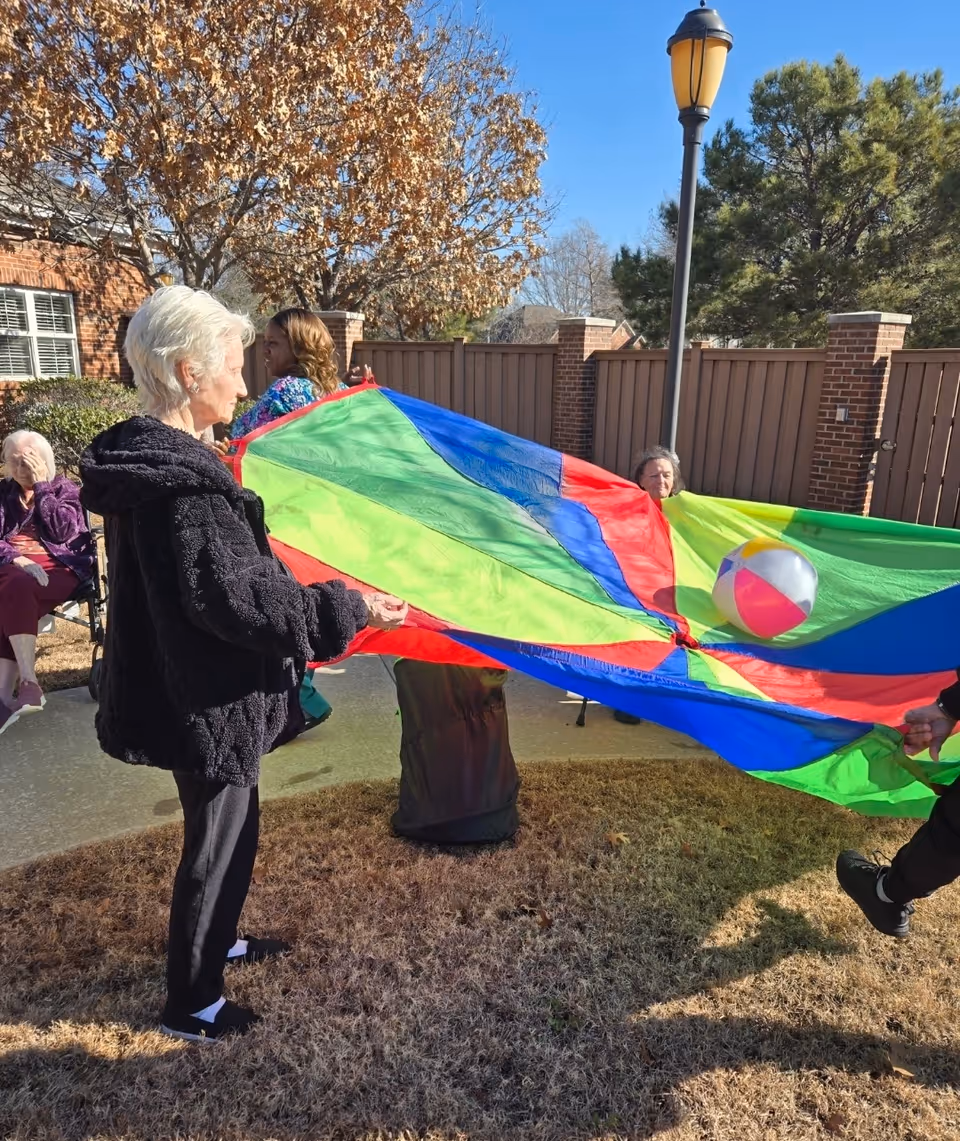 A group of elderly people outdoors holding the edges of a colorful parachute with a beach ball on top of it, surrounded by a brick fence and trees under a clear blue sky.