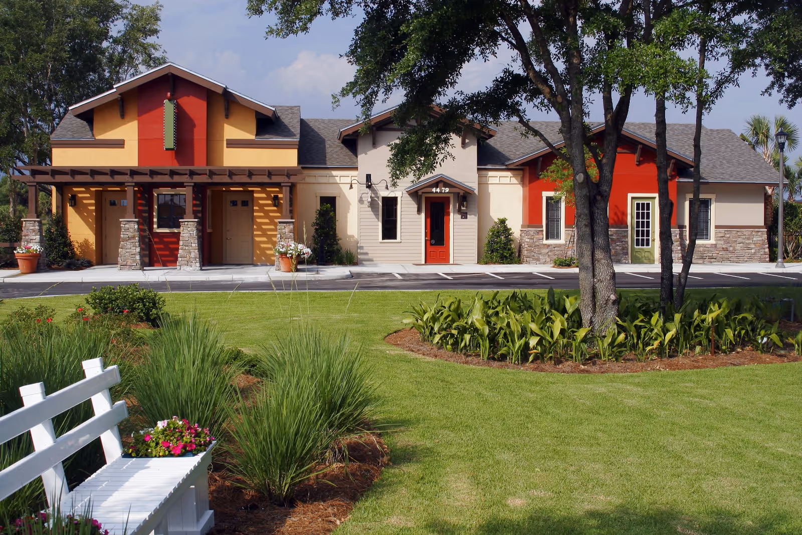 Exterior view of a colorful single-story building with red, yellow, and beige walls, stone accents, and multiple doors. In the foreground, there is a well-maintained lawn with green grass, landscaped plants, trees, and a white bench with a flower pot on it.