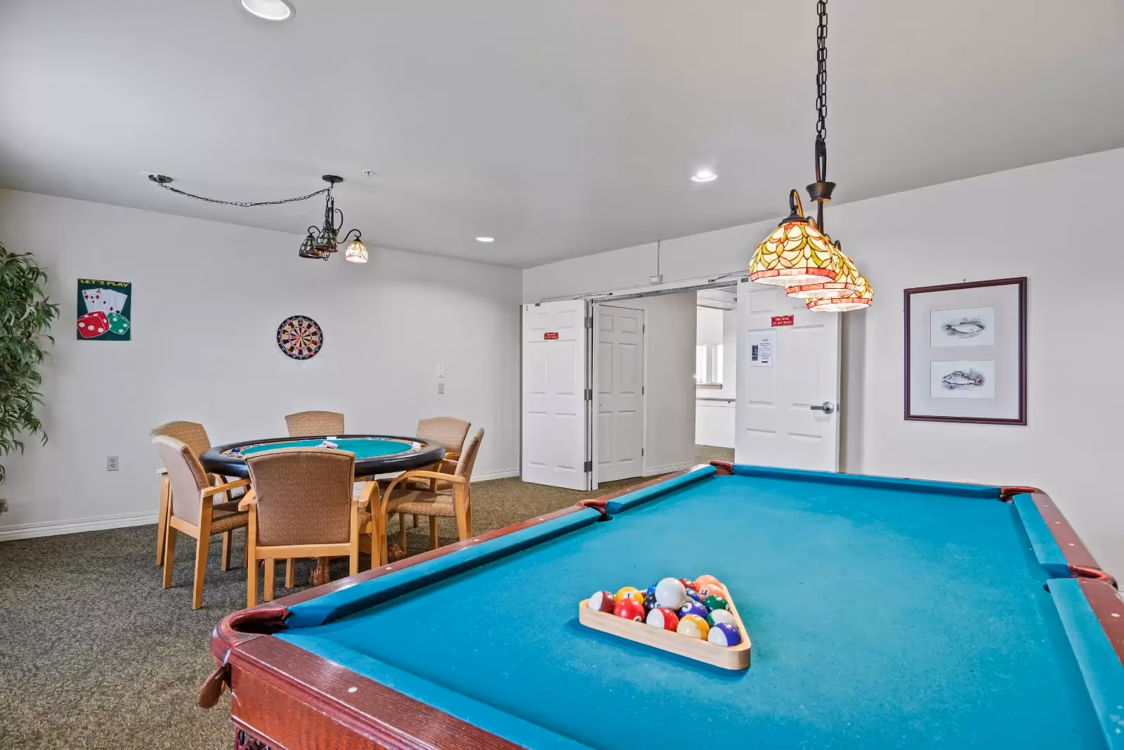 A game room with a pool table in the foreground, set up with billiard balls in a triangle rack. In the background, there is a round card table with four chairs around it, a dartboard on the wall, and a small sign with playing cards and dice. The room has white walls, carpeted floor, and two hanging stained glass pendant lights above the pool table. Double doors are open leading to another room.