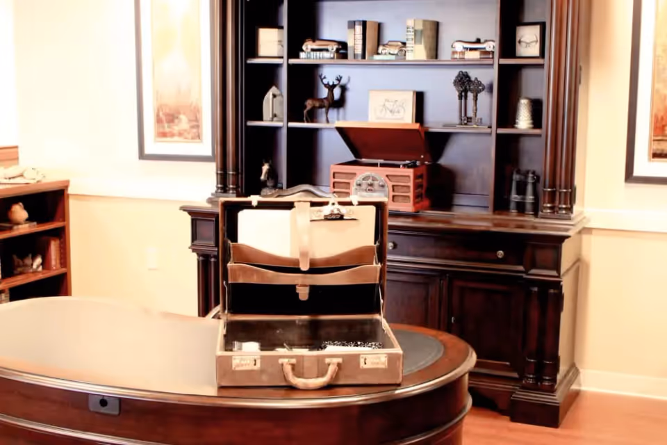 A warmly lit office or study room featuring a dark wooden desk with an open vintage briefcase on top. Behind the desk is a large dark wooden bookshelf with decorative items including books, small sculptures, and a vintage-style record player. The walls are light-colored with framed artwork on either side of the bookshelf.