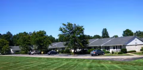 Single-story senior living building with cars parked along a curved driveway and manicured lawn.