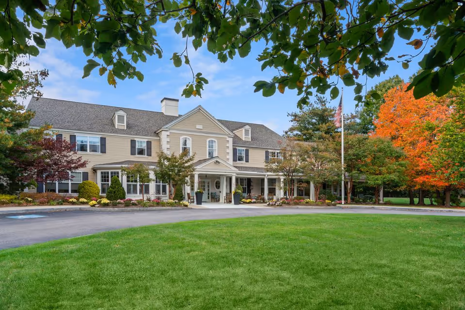 Exterior view of a two-story senior living facility building with beige siding, white trim, and a covered entrance. The building is surrounded by well-maintained landscaping including trees with green and autumn-colored leaves, shrubs, and flower beds. A green lawn and a paved driveway are visible in the foreground under a partly cloudy sky.
