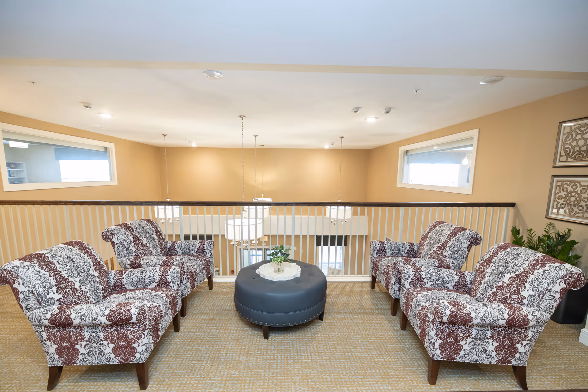 A cozy seating area in a senior living facility with four patterned armchairs arranged around a round black ottoman with a small plant on top. The area is carpeted and overlooks a lower level through white railing. The walls are painted beige with two small windows and decorative wall art on the right side.