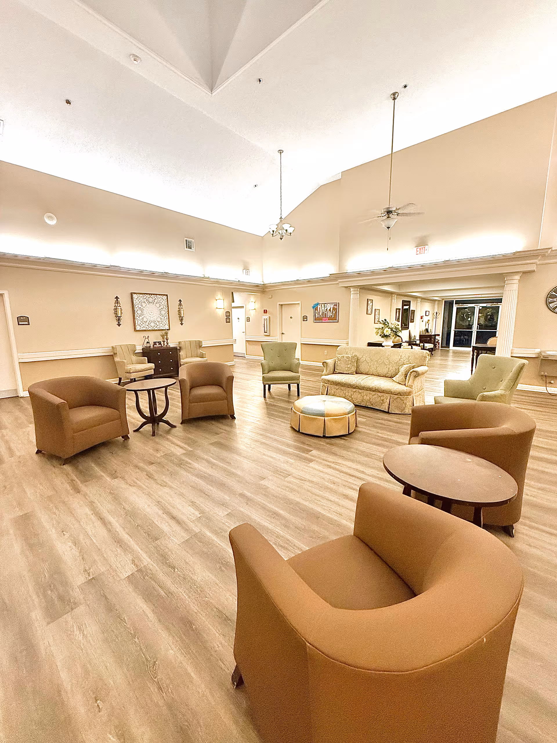 A spacious senior living facility common area with high ceilings and warm lighting. The room features several comfortable chairs arranged in a circle around small tables, a beige patterned sofa, and a round ottoman in the center. The walls are decorated with framed artwork and sconces, and there is a 'Happy Birthday' sign visible in the background near a hallway entrance.