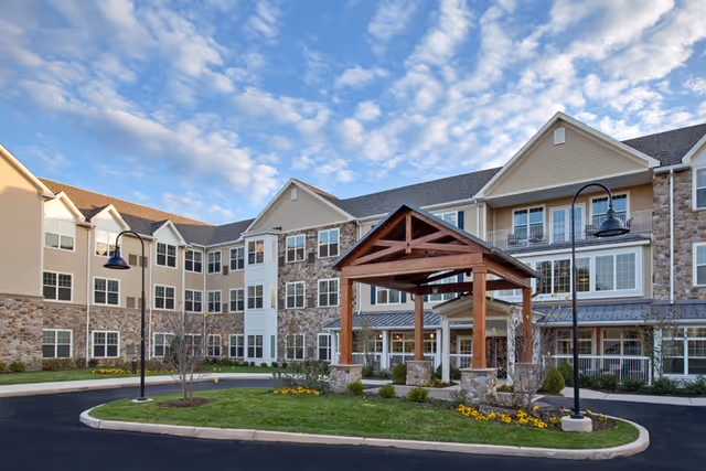 Exterior view of The Solana Doylestown senior living facility showing a three-story building with stone and beige siding, multiple windows, a covered entrance with wooden beams, landscaped greenery, and a paved driveway under a partly cloudy sky.
