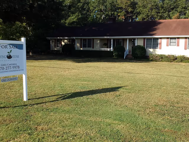 Single-story residential building with a large front lawn and a 'For Sale' sign in the foreground. The building has a dark roof, light-colored walls, and several windows with red shutters. Trees are visible in the background.