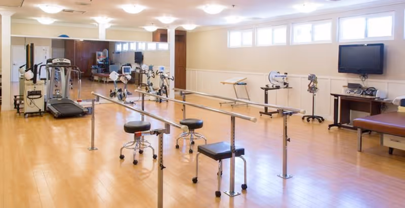 A spacious rehabilitation room with wooden flooring, parallel bars for walking exercises, several stools on wheels, exercise equipment including a treadmill, and a wall-mounted television. The room is well-lit with ceiling lights and windows near the ceiling.