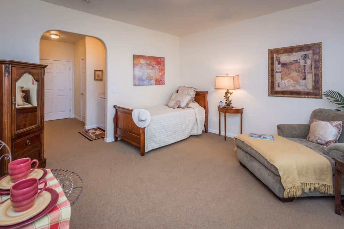 A cozy senior living room with a single wooden bed covered with a beige quilt and pillows, a wooden side table with a lamp, a cushioned chaise lounge with a yellow throw blanket and pillows, a wooden cabinet with a mirror, and a small table set with pink cups and plates. The walls are decorated with framed artwork, and there is a carpeted floor.