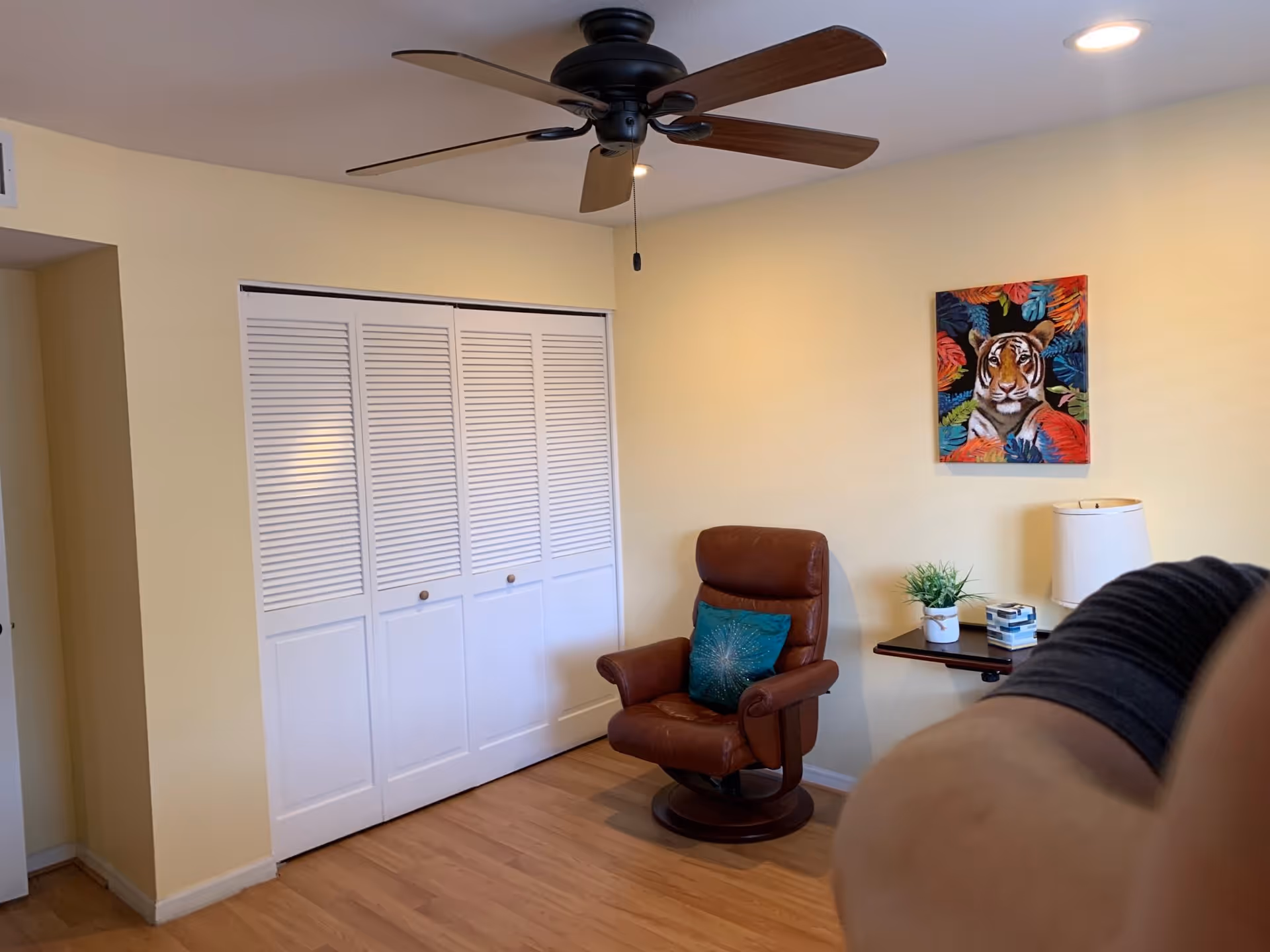 A cozy room with light yellow walls and wooden flooring. There is a brown leather armchair with a teal cushion, a small wall-mounted shelf holding a potted plant, a lamp, and some books. Above the shelf, a colorful painting of a tiger surrounded by tropical leaves is hung. A ceiling fan with wooden blades is mounted on the ceiling. White louvered closet doors are visible on one wall.